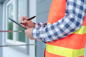 Close up of man in plaid shirt with an orange safety vest looking over a clipboard in front of a home