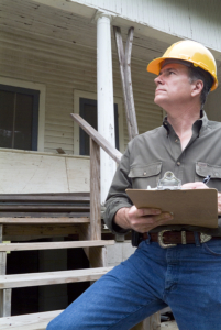 A man in a hard hat standing in front of an old rundown house holding a clipboard in his hand